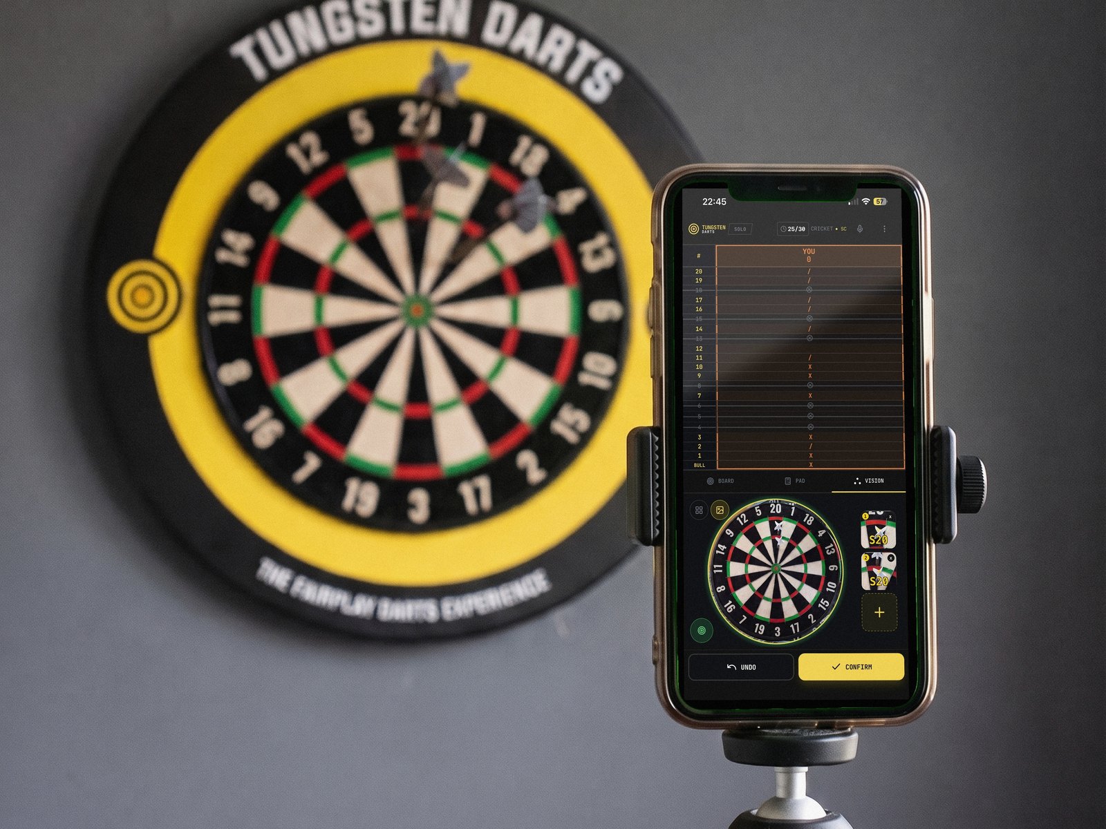 A phone on a small tripod in front of a Tungsten Darts dartboard, the app reading the darts and showing the score